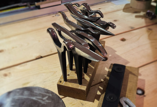 An array of metal forming tools, including hammers and anvils, neatly arranged on a wooden workbench in a home studio. The tools are used for crafting art jewelry from silver and copper at MorphiArtforge